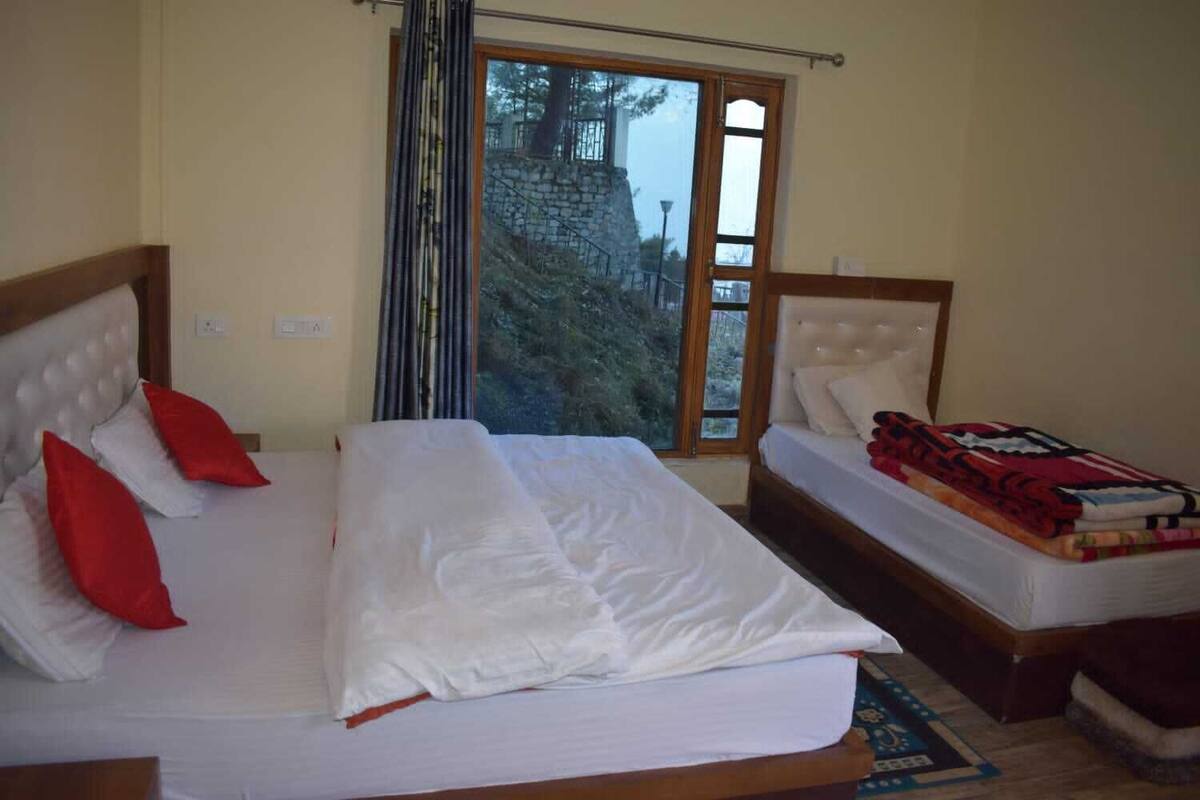 Una cama doble con ropa blanca y almohadas rojas, una cama más pequeña con ropa blanca, una ventana con vistas a árboles y un muro de piedra, y un cabecero de madera.
