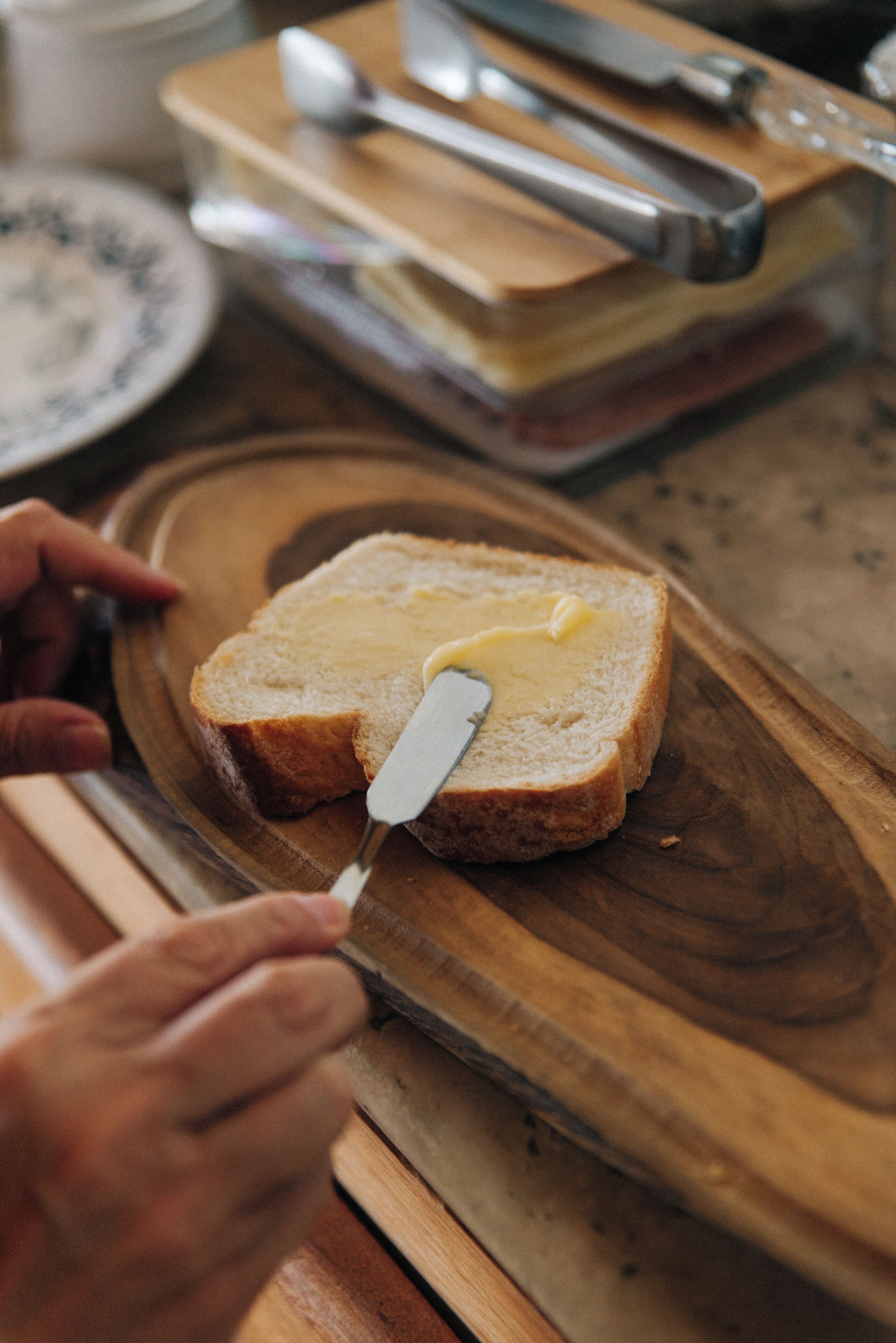 A wooden cutting board with a slice of bread and a knife spreading butter on it.