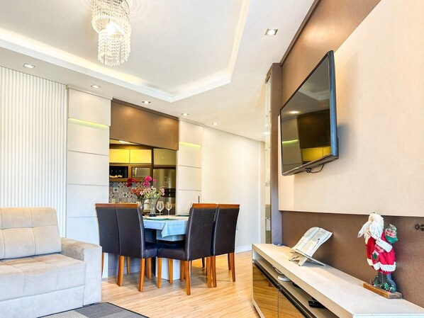 A modern living room with a dining area, a flat-screen TV mounted on the wall, and a small table with a Santa figurine.