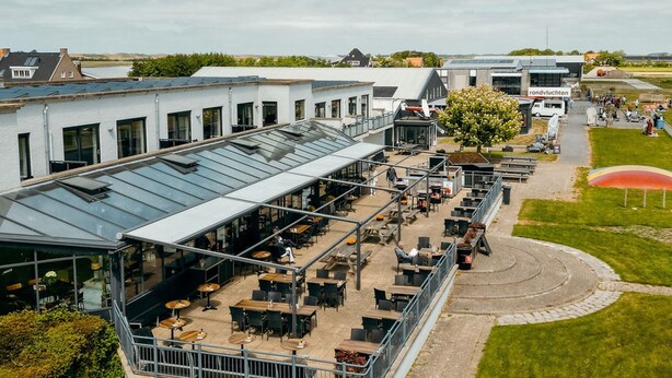A modern outdoor café with a glass roof, surrounded by neatly arranged tables and chairs, set against a backdrop of residential buildings and a clear sky.