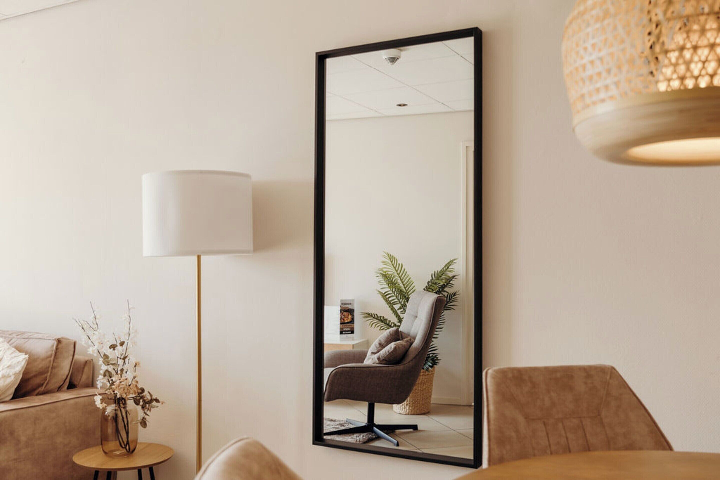 A modern living room with a large mirror, a sofa, a small round table with a vase, and a lamp.