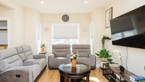 Ein Wohnzimmer mit grauem Sofa, grauem Sessel, Glastisch, Flachbildfernseher und Bücherregal mit Büchern und Pflanze.