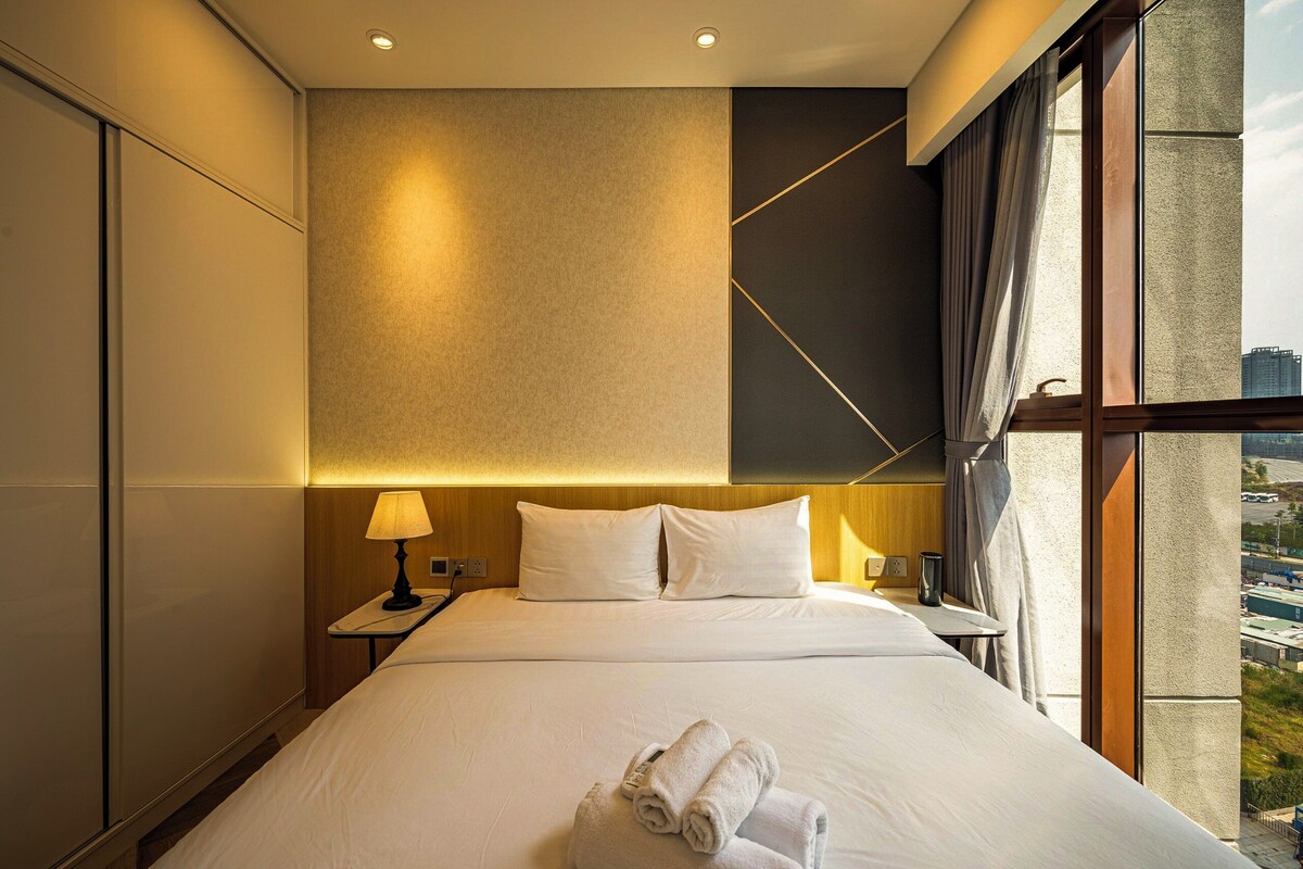 A hotel room with a large bed, a nightstand with a lamp, and a view of the city through the window.
