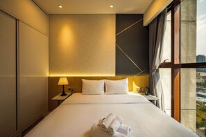 Habitación de hotel con una cama grande, una mesita de noche con lámpara y vistas a la ciudad desde la ventana.