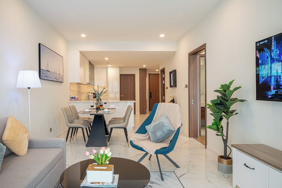 A modern living room with a sofa, a coffee table with flowers, a dining area with chairs, and a kitchen in the background.