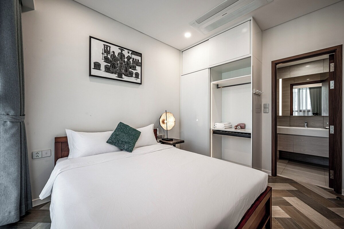 Una habitación de hotel moderna con una cama grande, una mesita de noche con lámpara, una obra de arte en la pared y un baño visible a través de una puerta abierta.