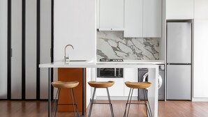 Een moderne keuken met een wit aanrecht, drie houten krukken, een inbouwoven en een roestvrijstalen koelkast.