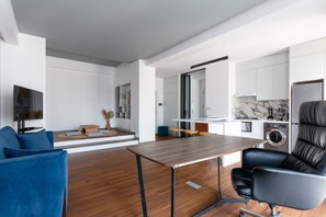 A modern living space with a wooden dining table, a blue sofa, a black leather chair, and a kitchen area with white cabinets and a marble backsplash. - Boulevard 114 (Tashkent)