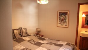 Una camera da letto con un letto, un comodino, una lampada, un poster appeso al muro e un bagno con lavandino e specchio.