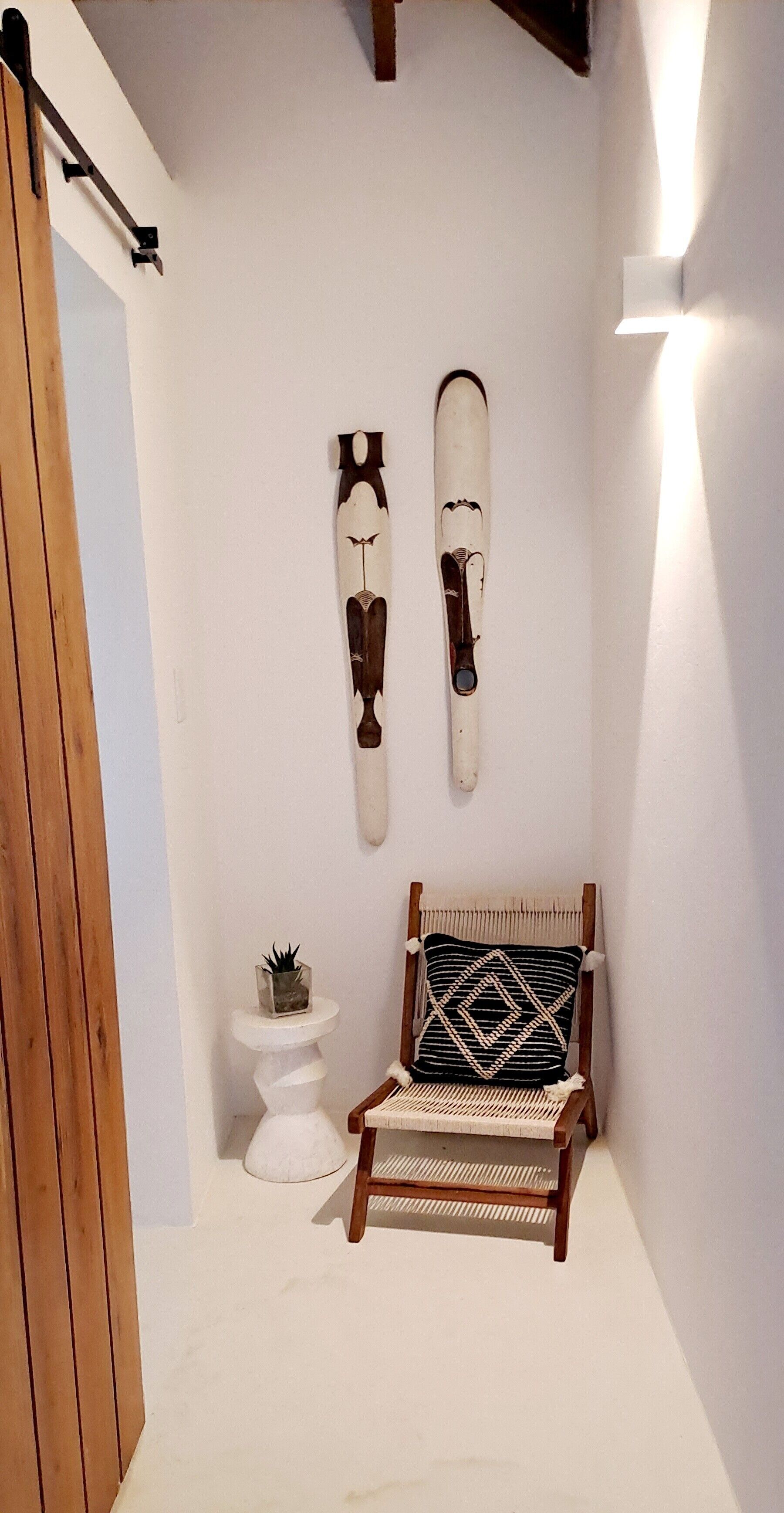 A small, cozy room with a wooden chair, a white pedestal table with a plant, and two wall-mounted decorative items.