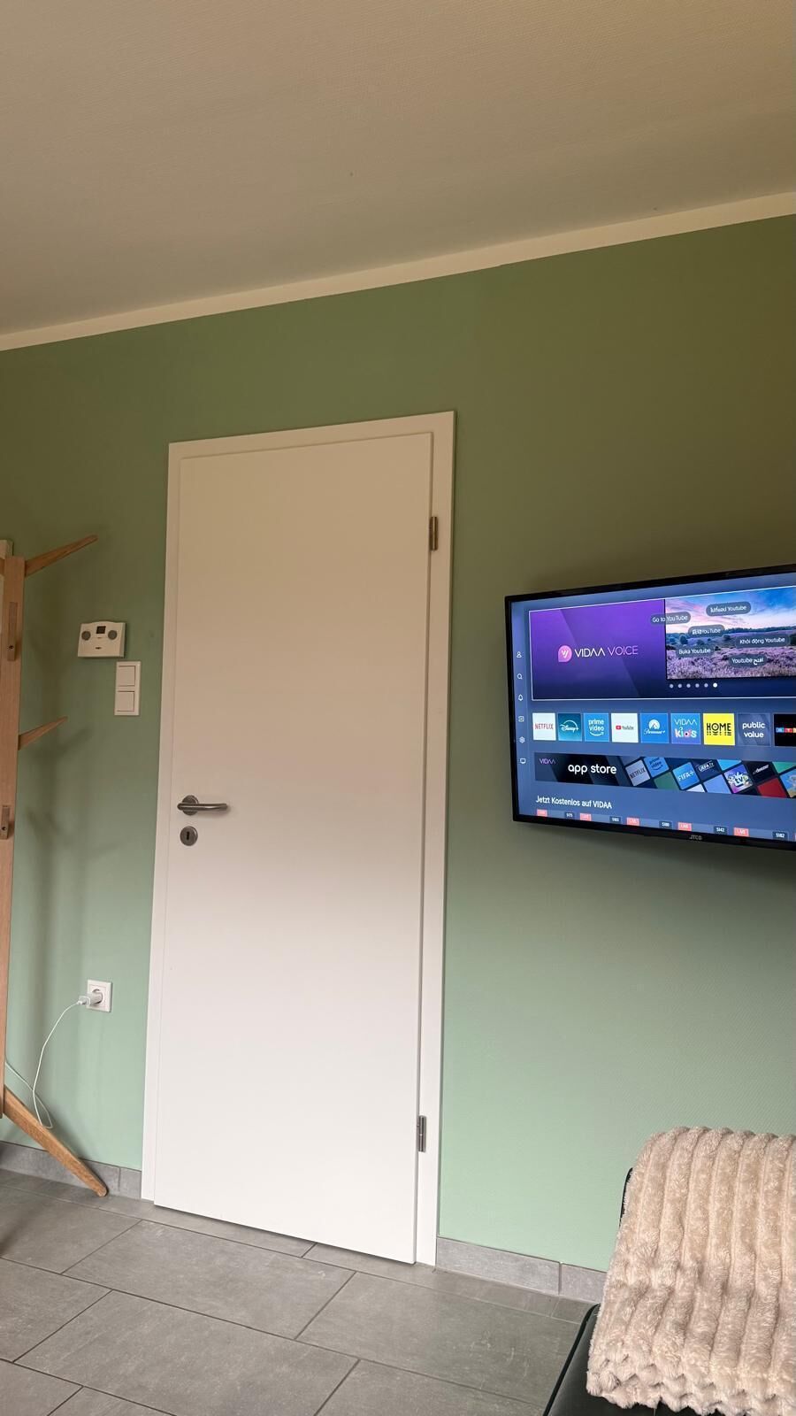 Une pièce avec une porte blanche, un téléviseur fixé au mur qui affiche l’interface d’un service de diffusion en continu, et un sol carrelé.