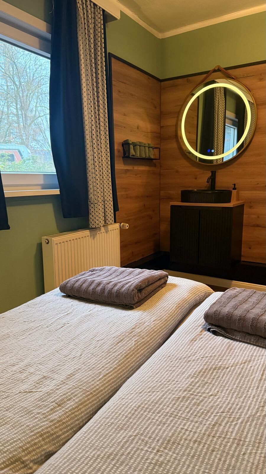 Ein Zimmer mit zwei Betten, einem runden Spiegel mit Beleuchtung, einem Heizkörper und einem Fenster mit Vorhängen.