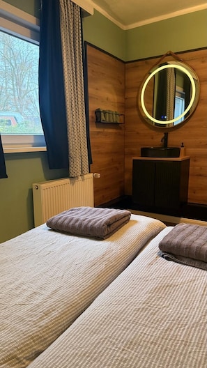 A room with two beds, a round mirror with a light, a radiator, and a window with curtains. - Cozy bungalow in a vacation complex in the Teutoburg Forest. (Lage)