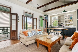A spacious living room with a wooden coffee table, a beige sofa with patterned cushions, and a kitchen area with bar stools and shelves of kitchenware. - The Blue Chip | Large Veranda | Al Fresco Dining (Charleston)