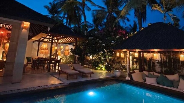 A pool area with lounge chairs and a covered seating area, surrounded by palm trees and a night sky. - VILLA DUNIA (Gili Trawangan)