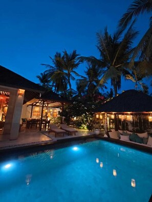 A pool area with lounge chairs and a covered seating area, surrounded by palm trees and a night sky. - VILLA DUNIA (Gili Trawangan)