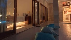 A modern hotel room with a large sliding glass door leading to a balcony, a bed with a nightstand, and a small table with a lamp. - VILLA DUNIA (Gili Trawangan)