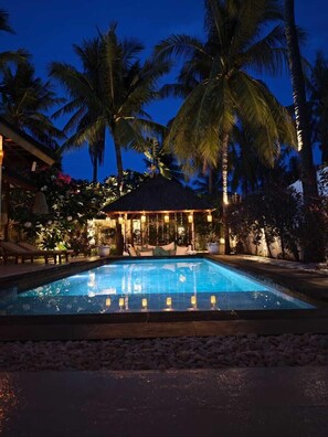 A pool area with a lounge chair and a covered seating area surrounded by palm trees. - VILLA DUNIA (Gili Trawangan)