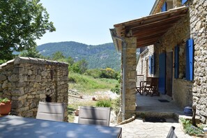 Ein Steinhaus mit überdachter Terrasse, Sitzgelegenheiten im Freien und Blick auf eine gebirgige Landschaft.