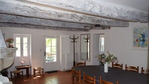 Comedor con una mesa grande, sillas de madera y una araña. Hay ventanas con cortinas, un radiador y un florero con flores sobre una mesita.