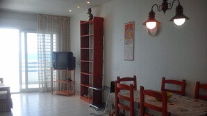 A living room with a television on a stand, a dining table with chairs, a bookshelf, and a large window with curtains. - First Line Apartment With Wifi (Benicarlo)