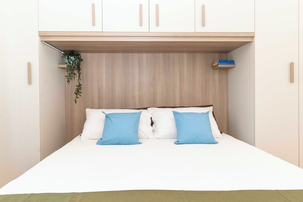 A bed with white bedding and two blue pillows, a wooden headboard, and a small shelf with a plant.