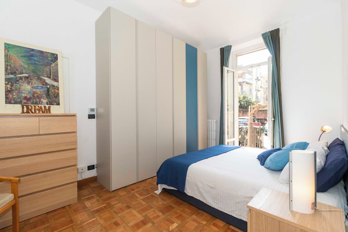A bedroom with a bed, a wardrobe, a desk, and a window with curtains.
