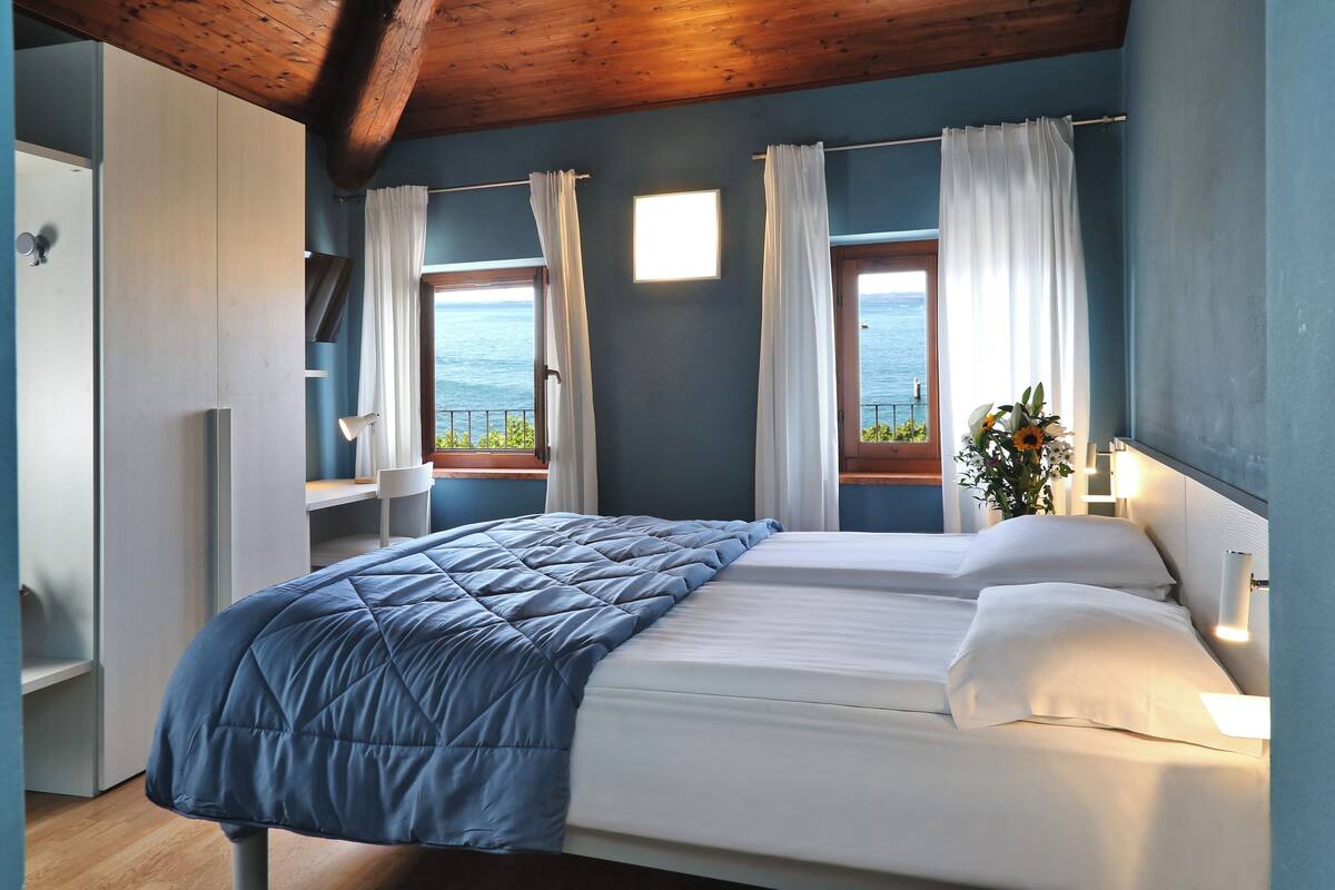 Quarto com cama, duas janelas com cortinas, escrivaninha, cadeira e vista para o mar.