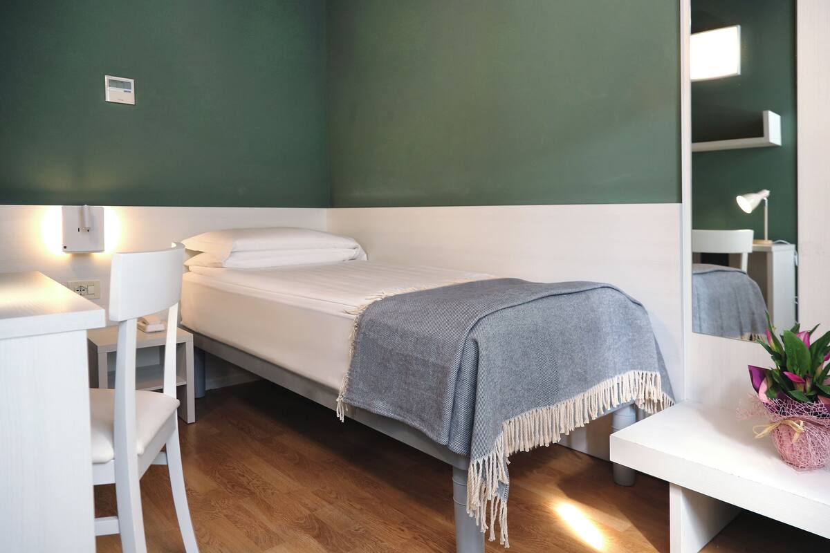 Quarto pequeno e bem organizado, com uma cama de solteiro, uma escrivaninha, uma cadeira e uma mesa de cabeceira com abajur e planta em vaso.