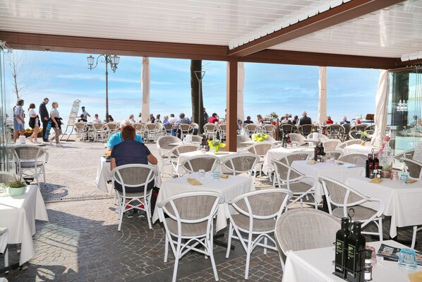 An outdoor restaurant with white wicker chairs and tables, a clear sky, and a view of the sea.