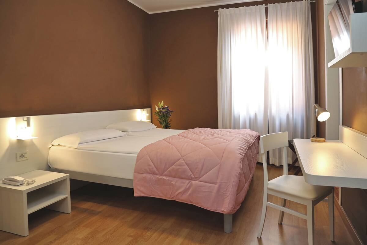 Quarto bem arrumado com cama, mesa de cabeceira, cadeira e escrivaninha com luminária.