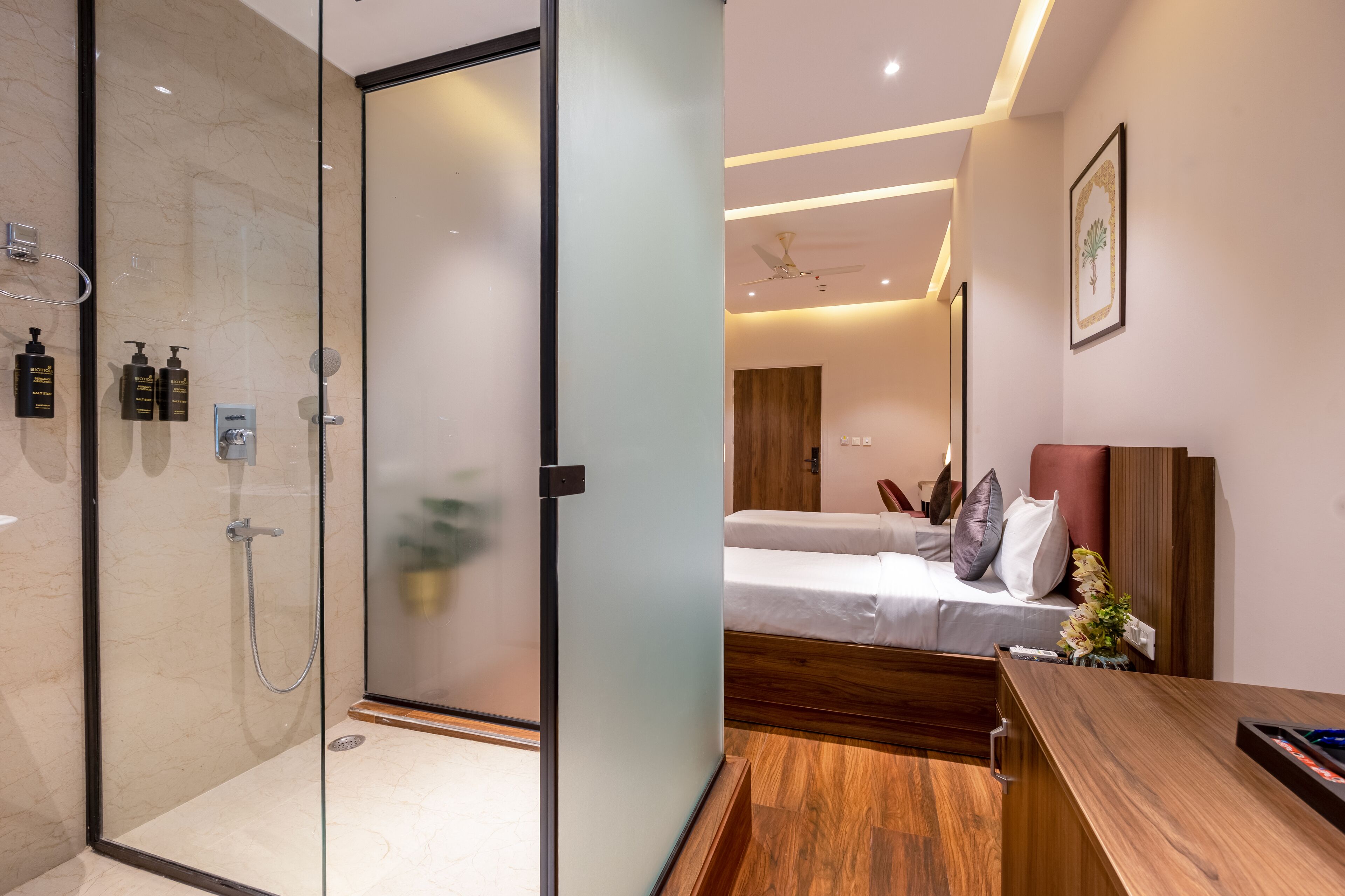 A modern hotel room with a glass-enclosed shower, a wooden bed with white linens, and a wooden desk with a vase of flowers.