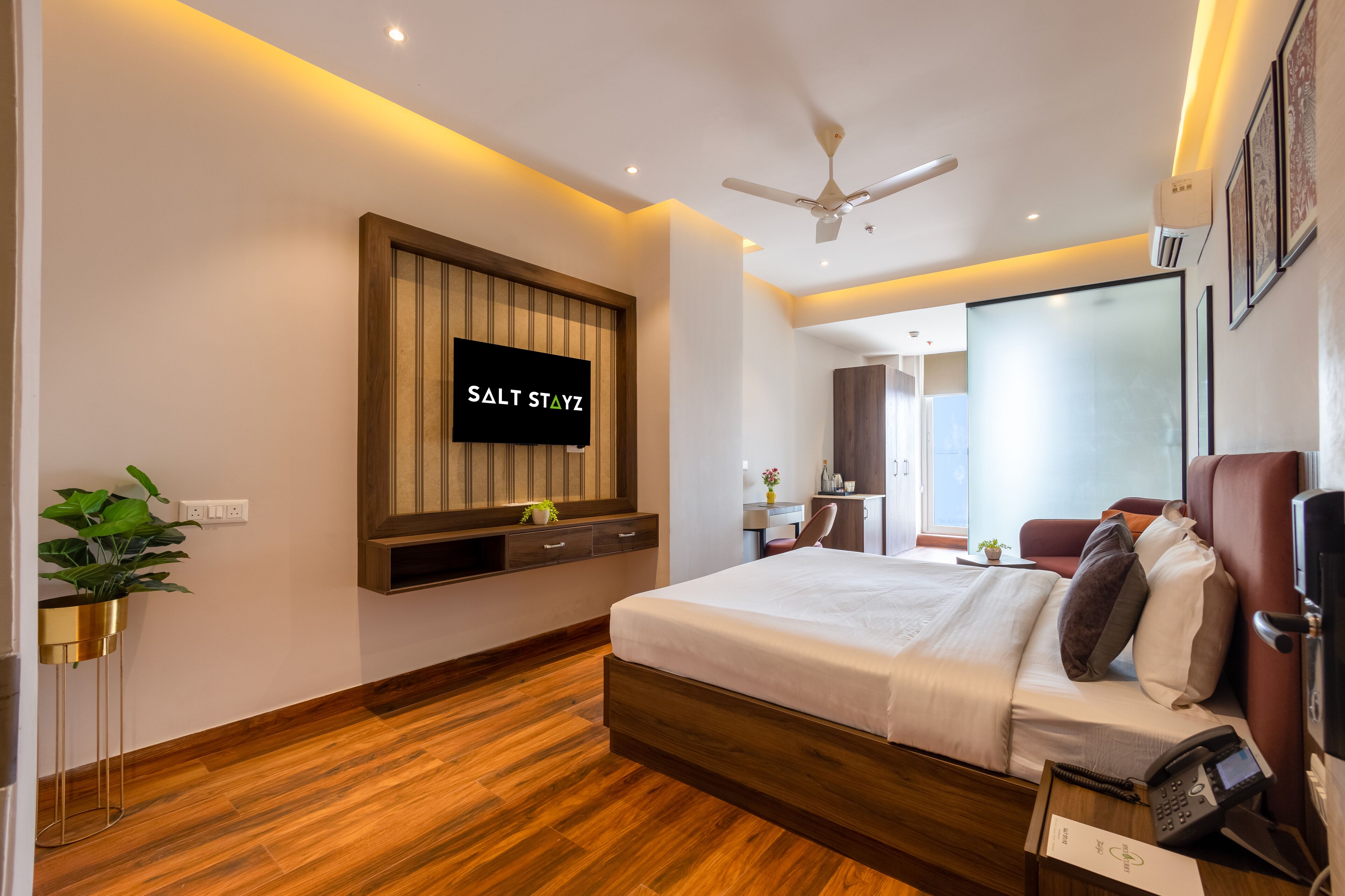 A modern hotel room with a large bed, a mounted TV, a ceiling fan, and a wooden floor.
