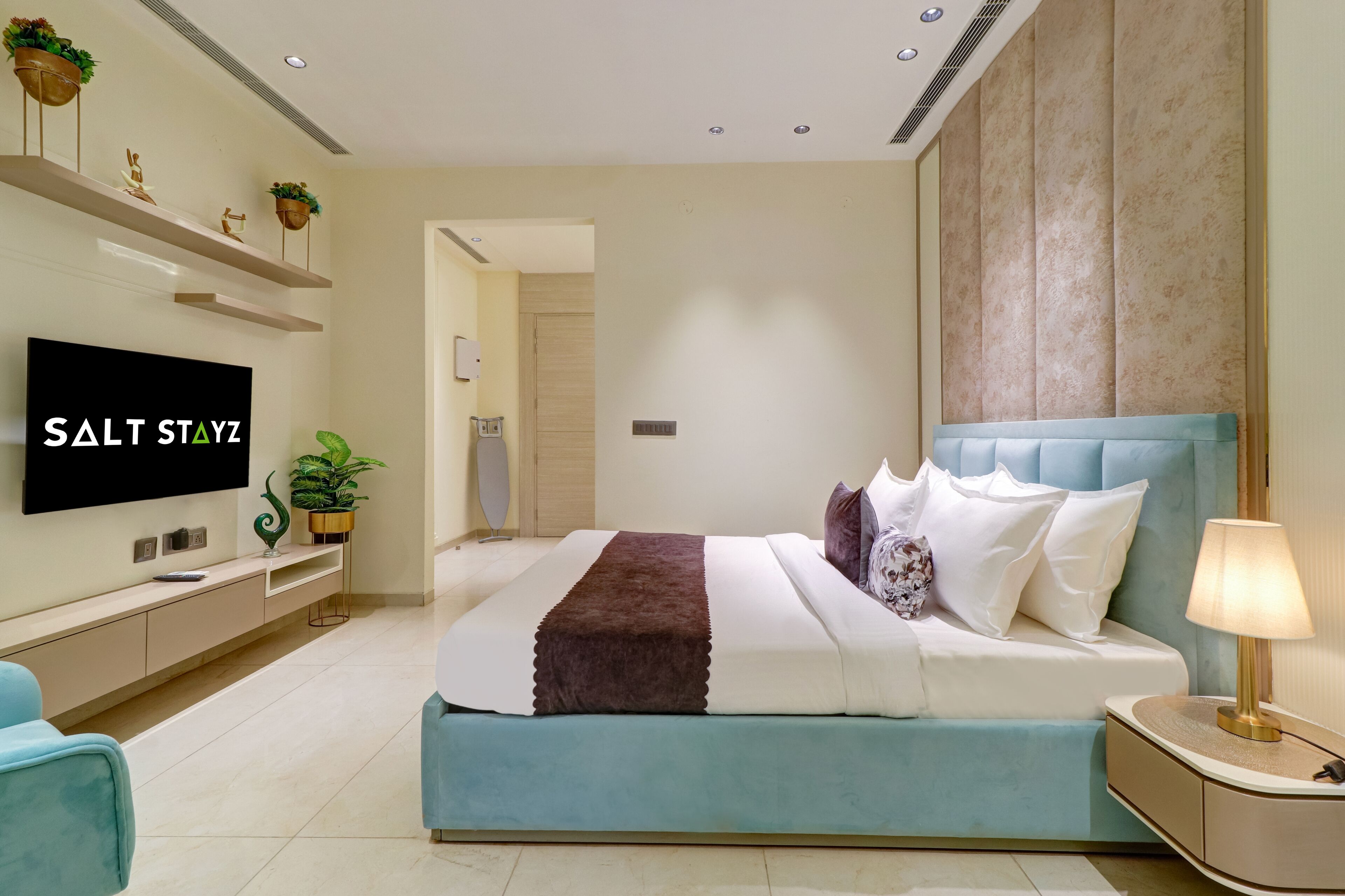A modern bedroom with a large bed, a TV, a bedside table with a lamp, and a wall-mounted shelf with decorative items.
