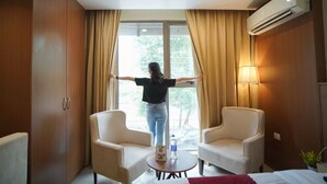 Une chambre d’hôtel avec une grande fenêtre, deux fauteuils, une petite table avec une bouteille et un appareil de climatisation.