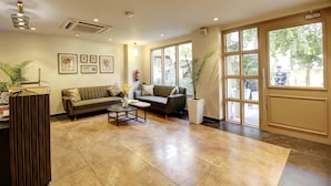 A spacious living area with a sofa, coffee table, and potted plants. There are large windows allowing natural light and views of the outside. - Saltstayz Select - Golf Course Road & Sector 27 (Gurugram)