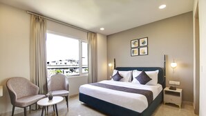 A hotel room with a large bed, two armchairs, a small table with a plant, a nightstand with a lamp, and a window with curtains. - Saltstayz Select - Golf Course Road & Sector 27 (Gurugram)