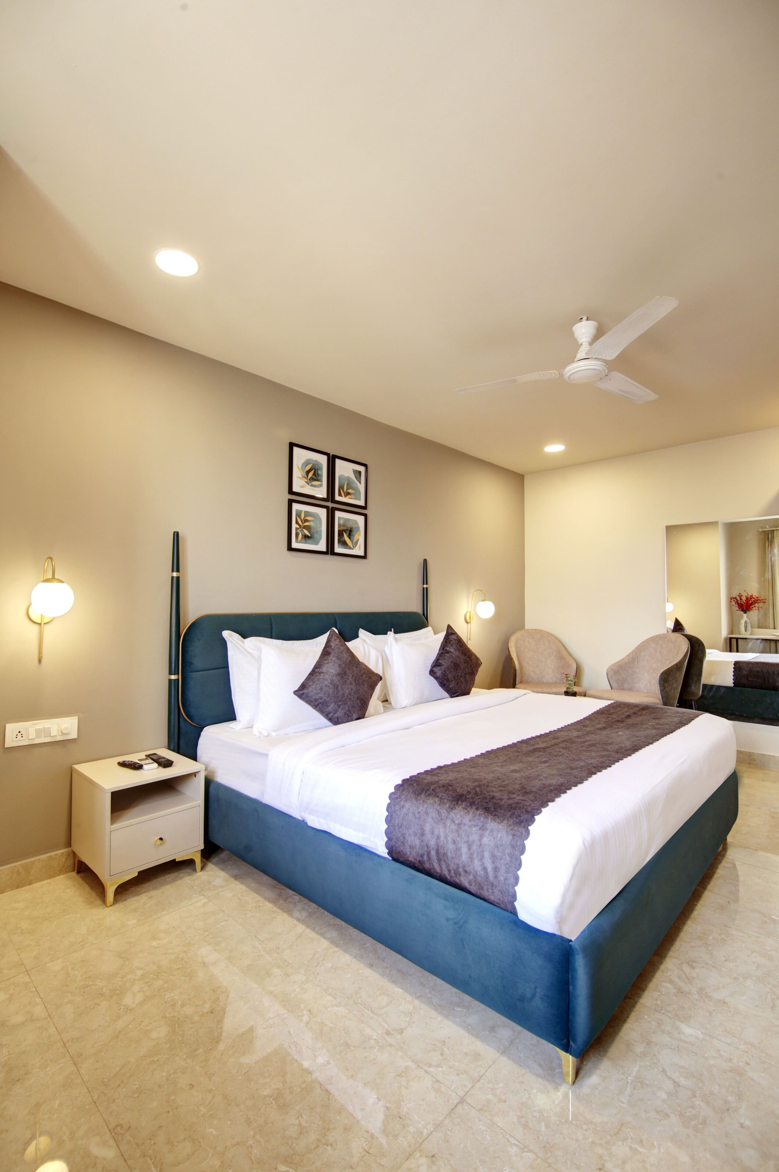 Une chambre d’hôtel bien aménagée, avec un grand lit, une table de chevet, une chaise, un ventilateur de plafond et des œuvres d’art accrochées au mur.