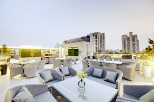 Una terraza en la azotea con mesas al aire libre, una zona de bar y vistas a edificios altos.