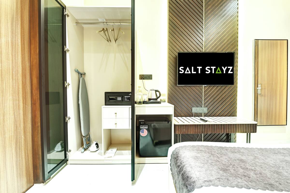 Ein modernes Hotelzimmer mit einer Schiebetür zu einem Duschbereich, einem kleinen Schreibtisch mit Computer, einem an der Wand montierten Fernseher und einem Bett mit grauer Decke.