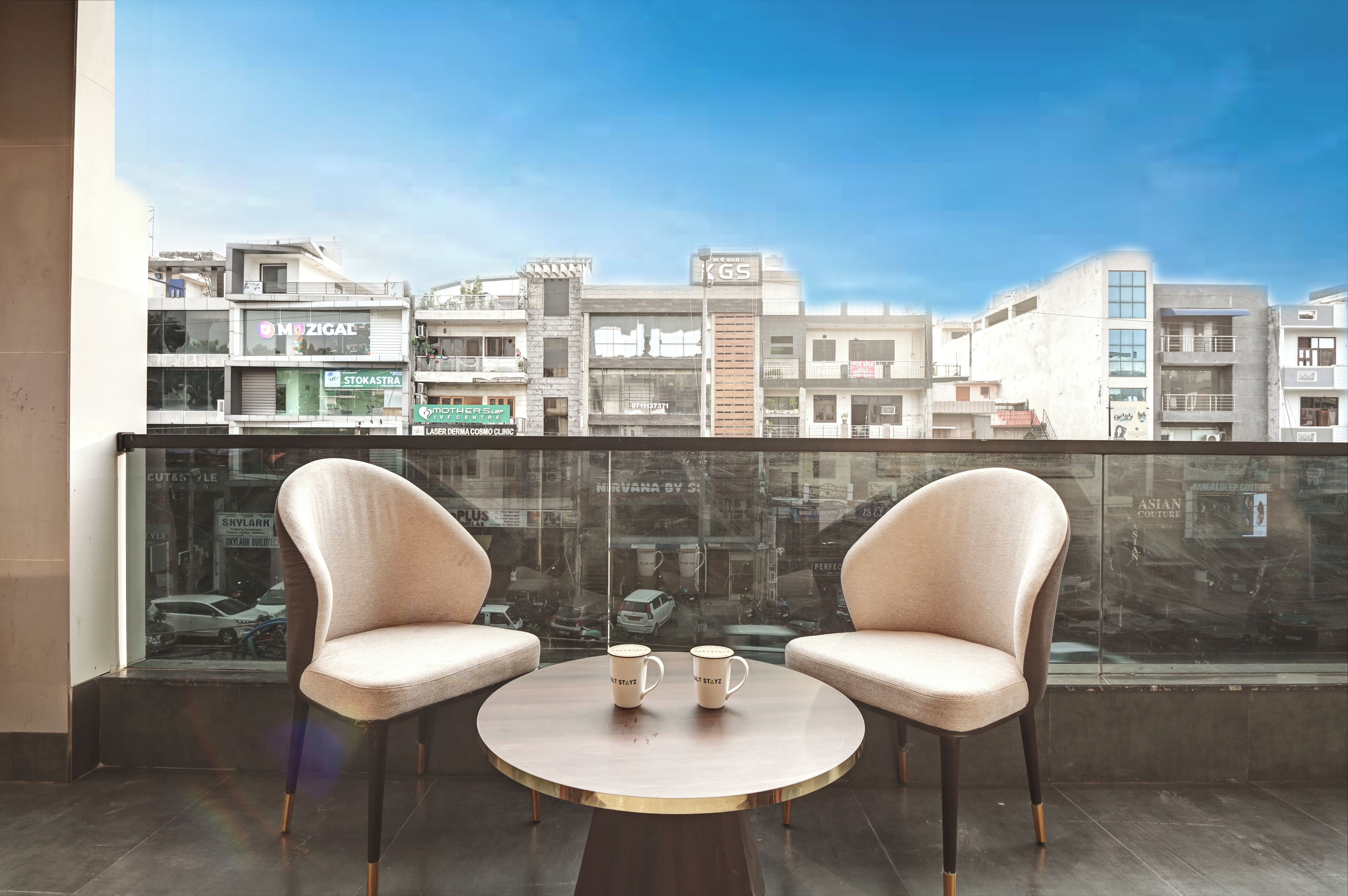 A balcony with two chairs and a table, overlooking a cityscape with various buildings and shops.