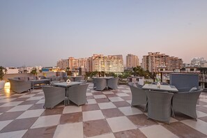 A rooftop terrace with checkered flooring, wicker furniture, and a cityscape view in the background. - Saltstayz Premier – Golf Course Road & Sector 42 (Gurugram)