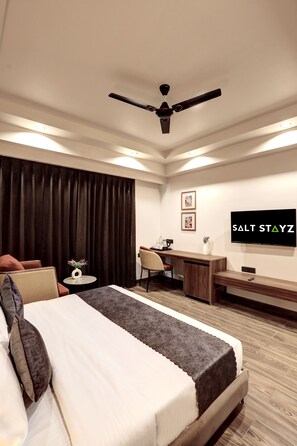 A hotel room with a bed, desk, chair, TV, and curtains. - Saltstayz Premier – Golf Course Road & Sector 42 (Gurugram)