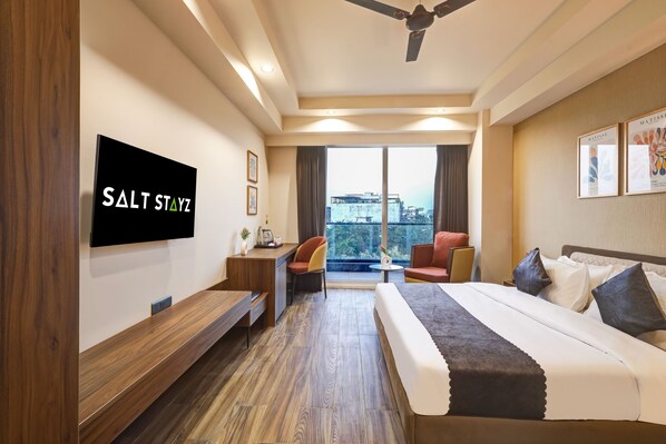 A hotel room with a bed, a desk, a chair, a TV, and a balcony with a city view. - Saltstayz Premier – Golf Course Road & Sector 42 (Gurugram)