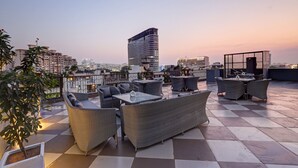 A rooftop terrace with wicker furniture, a glass table, and a view of a cityscape at dusk. - Saltstayz Premier – Golf Course Road & Sector 42 (Gurugram)