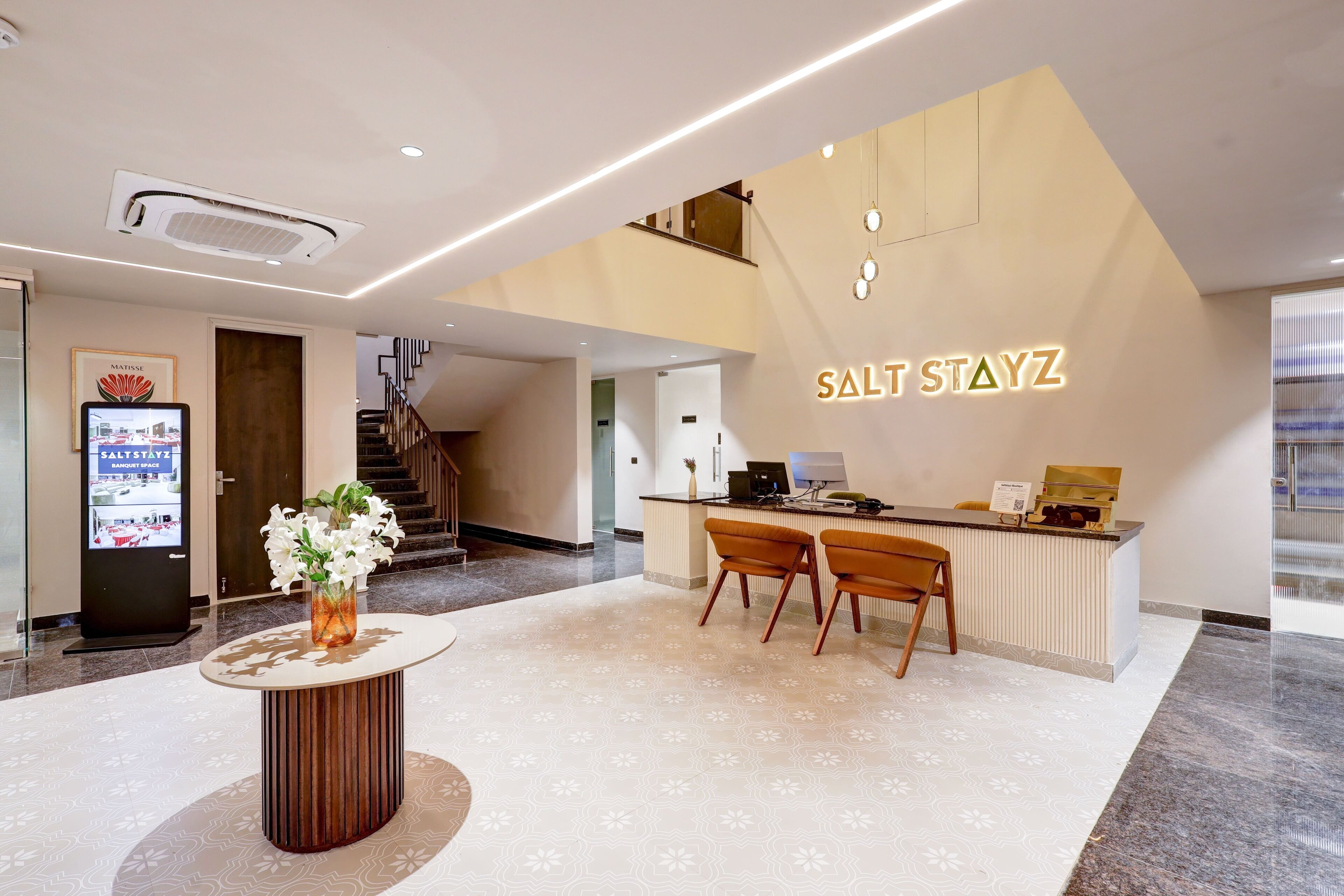 A hotel reception area with a modern design, featuring a reception desk, two chairs, a vase with flowers, and a digital display board.