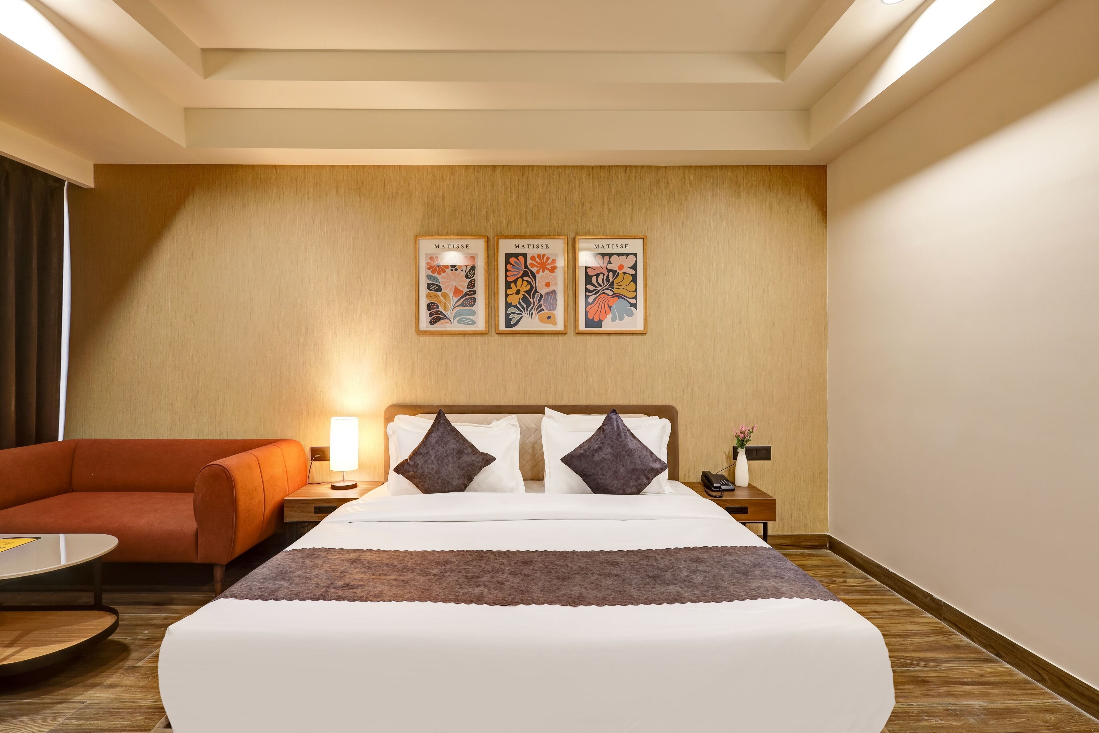 A hotel room with a bed, a sofa, a nightstand, a lamp, and three framed artworks on the wall.