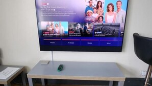 Un televisor de pantalla plana montado en una pared sobre un piso de madera, con un control remoto en una mesita auxiliar a un lado.