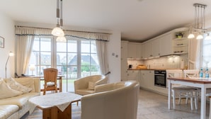 Eine moderne Küche mit weißen Schränken, eine Essecke mit Holztisch und -stühlen sowie ein Wohnzimmer mit Sofa und Couchtisch.