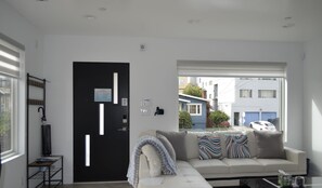 Ein modernes Wohnzimmer mit einer schwarzen Tür, einem Sofa mit gemusterten Kissen, einem Couchtisch und Blick auf eine Straße mit Gebäuden und parkenden Autos.